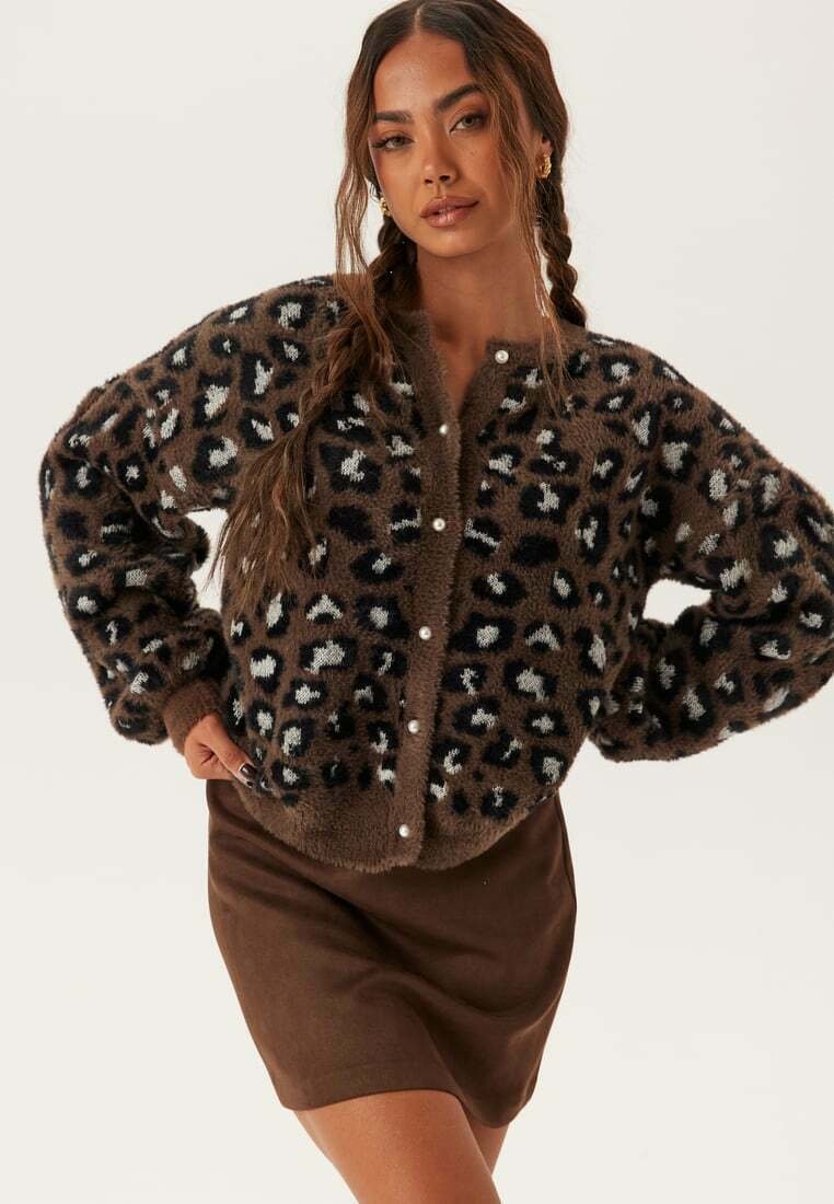 Kvinna med flätat hår som bär en brun leopardmönstrad fluffig cardigan med pärlknappar och en matchande brun kjol, poserar med händerna på höfterna.