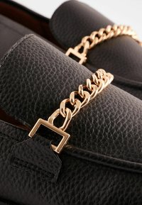 Black leather loafers featuring a textured finish and a gold chain accent across the upper. Square hardware detail near the base of the chain.