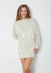 Ugličasta haljina u boji off-white, s opuštenim krojem, dugim prozirnim rukavima i strukiranim pojasom. Glatka tekstura s reflektirajućim šljokicama.