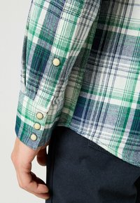 Koszula w kratę w kolorze zielonym i niebieskim na białym tle, z guziczkami na mankietach i gładką teksturą materiału.