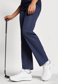 Navy golf pants, relaxed fit, paired with white athletic shoes. A golf club held in hand accents the sporty outfit.