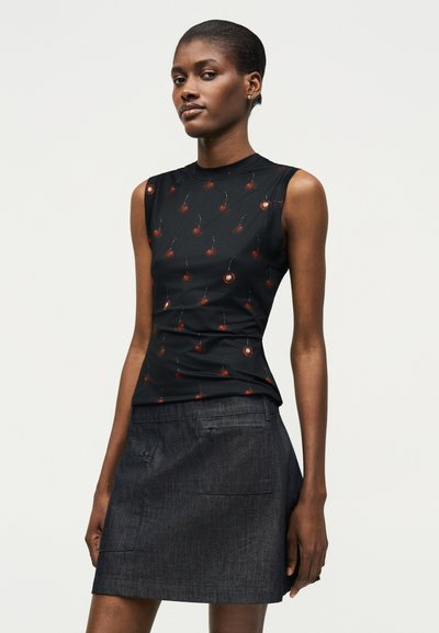 Γυναίκα με μαύρο αμάνικο τοπ με σχέδιο κεράσια και σκουρόχρωμη γκρι τζιν φούστα, στέκεται μπροστά σε απλό φωτεινό φόντο.