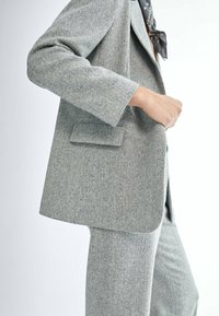 Osoba ubrana w jasnoszary garnitur w rybią kratę ze współmatchingową marynarką i spodniami, uzupełniona o ciemny szalik związany wokół szyi.