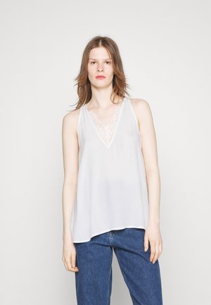 Hvid ærmeløs top med v-udskæring og blonder detaljer ved halsudskæringen, lavet af et let materiale, båret over blå denim jeans.