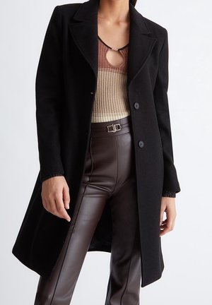 Kobieta w czarnym długim płaszczu na beżowo-brązowym prążkowanym topie z wycięciem przy dekolcie, w połączeniu z wysokim stanem ciemnobrązowych skórzanych spodni.