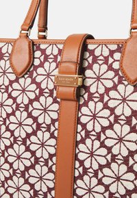 Braune Lederriemen sind an einer bordeauxroten Stofftasche befestigt, die ein weißes Blumenmuster aufweist. Ein goldenes Etikett trägt die Aufschrift "kate spade NEW YORK."