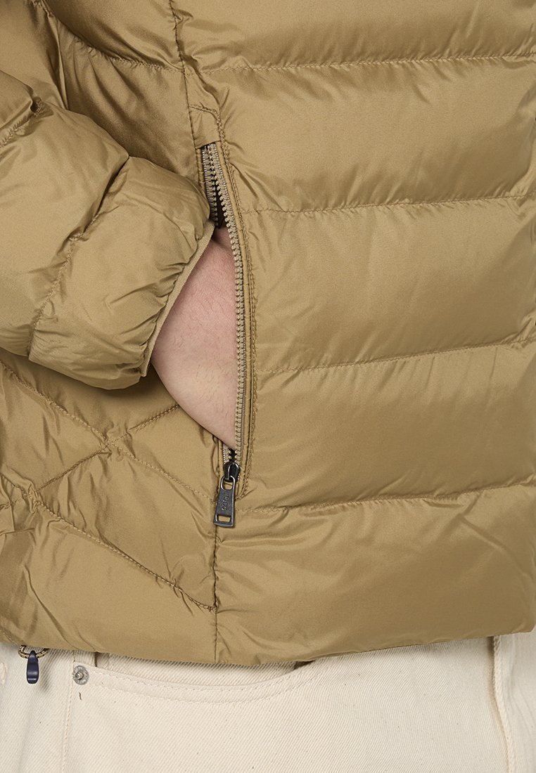 Beige Puffer-Jacke mit einem gesteppten Design und seitlichem Reißverschluss. Sie zeichnet sich durch eine leichte, glatte Textur und sichtbare Nähte aus.