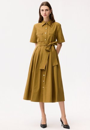 Vrouw draagt een midi-jurk met knopen in mosterdgeel, korte mouwen, een gebonden ceintuur in de taille en zwarte hoge hakken, staand tegen een effen witte achtergrond.