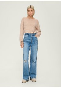 Maglietta a righe rosa chiaro con maniche lunghe abbinata a jeans blu a vita alta con dettagli strappati e gamba larga. Scarpe beige completano l'outfit.