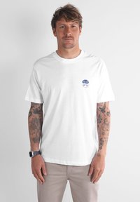 T-shirt en coton blanc à manches courtes et col rond. Présente un logo circulaire bleu sur la poitrine gauche. Texture lisse, coupe décontractée.