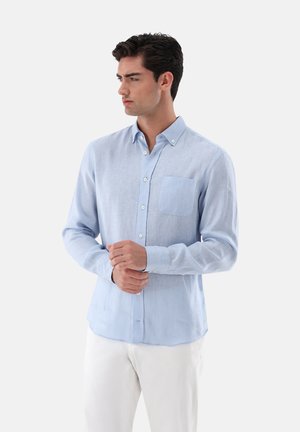 Hellblaues Langarmhemd aus strukturiertem Stoff, mit Button-Down-Kragen, Brusttasche und weißen Knöpfen. Getragen mit weißen Hosen.