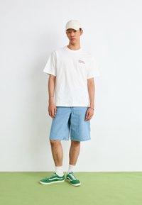 Homme portant une casquette blanche, un t-shirt blanc avec un petit texte, un short en denim bleu clair, des chaussettes blanches et des baskets vertes, debout sur un sol vert face à un mur blanc.
