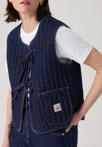 Dunkelblauer Denim-Weste mit vertikaler Stepptextur, Frontbindungen und einer aufgesetzten Tasche mit einem Logopatch. Über einem weißen T-Shirt getragen.