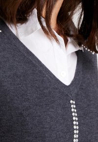 Grå stickad tröja med V-ringning, dekorerad med en rad klara pärlor längs mitten och lager-på-lager över en vit skjorta med knappar.