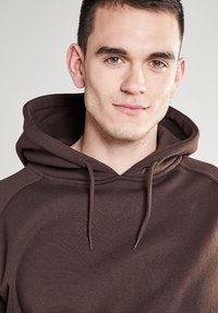 Felpa marrone realizzata in un materiale morbido, con cappuccio regolabile e dettagli a costine. Il design ha una vestibilità rilassata e cuciture piatte.