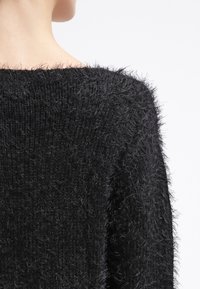 Suéter negro y peludo con un amplio escote al hombro y textura suave. La tela tiene un ligero brillo y un patrón de punto irregular.