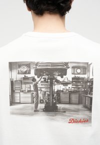 Μηχανικός εργάζεται κάτω από ένα αυτοκίνητο που έχει ανυψωθεί με υδραυλικό ανυψωτικό σε ένα συνεργείο γεμάτο με εργαλεία και ντουλάπια, με ένα ρολόι και μια σημαία ορατά.