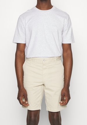 Homme portant un t-shirt à col rond gris clair et des shorts beige au genou, se tenant avec les bras détendus le long du corps.