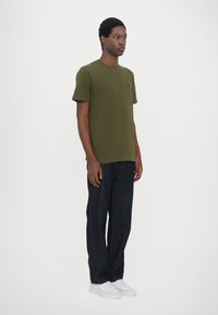 Tricou din bumbac de culoare verde măsliniu, cu mâneci scurte, guler rotund și un mic logo negru pe piept, asortat cu pantaloni din denim de un albastru închis și adidași albi.