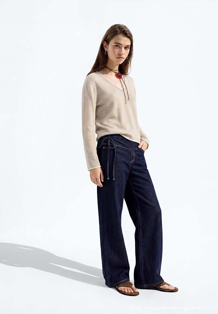 Jeune femme debout portant un pull beige à manches longues, un jean large foncé avec un détail noué, des sandales marron et un collier à fleurs rouges.