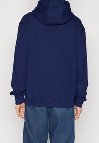 Tmavě modrá mikina s uvolněným střihem, velkou kapucí a žebrovanými manžetami. Měkká látka a minimální detaily, zkombinováno s volnými džíny.