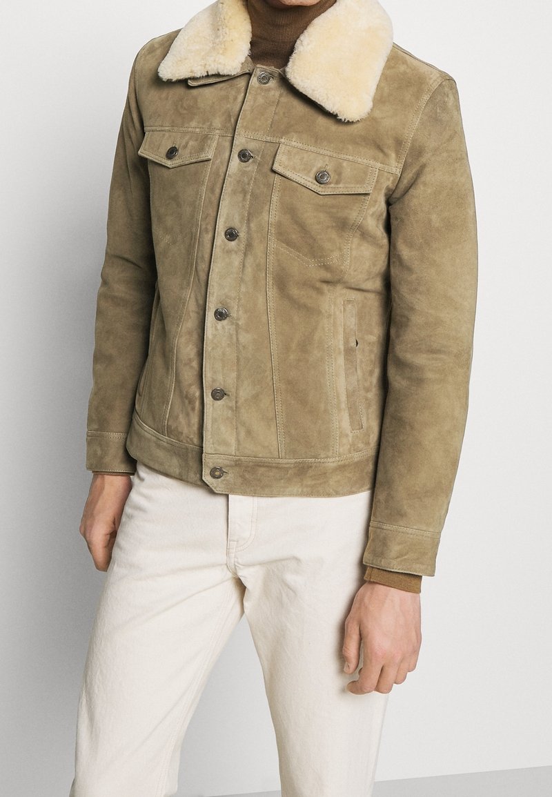 Béžová semišová bunda s plyšovým béžovým límcem z beránčí kožešiny, zapínáním na knoflíky vpředu a dvěma náprsními kapsami, charakterizovaná hladkou strukturou a střihovou úpravou na míru.