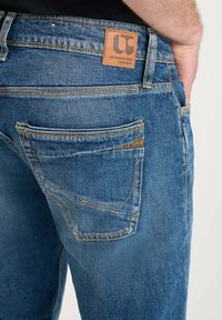 Jeans in denim blu con una patch in pelle etichettata "LE TEMPS DES CERISES" sul retro. Presentano tasche cucite e una texture scolorita.