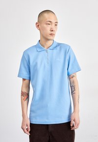 Polo shirt azul claro com fecho éclair, mangas curtas, tecido texturizado e logótipo bordado discreto. Ajuste casual com calças de veludo cotelê escuras.