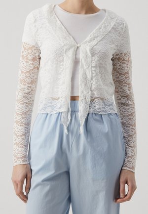 Femme portant un chemisier en dentelle florale blanche à manches longues sur un crop top blanc côtelé et un pantalon ample bleu clair.