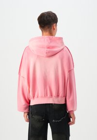 Pink hættebluse med sænkede skuldre, tætsiddende kant og blød tekstur. Sammen med slidte sorte jeans. Minimalistisk design.