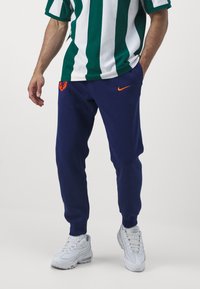 Námořnické tepláky s oranžovým logem Nike, se širokým elastickým pasem a manžetami. V kombinaci se zeleným a bílým pruhovaným tričkem.