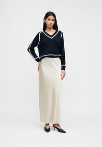 Σκούτερ μπλε ναυτικού με λευκή επένδυση, λαιμόκοψη V και μακριά μανίκια, σε συνδυασμό με μακριά, εκρού φούστα και μαύρες γόβες με μυτερή μύτη.