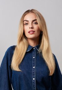 Chemise en denim avec col, fermeture à boutons sur le devant et manches longues. Présente une couleur bleue et un tissu texturé. Le mannequin a les cheveux longs et droits.