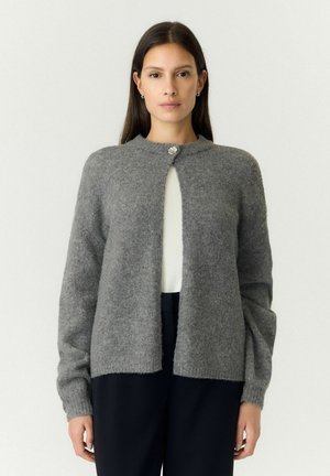 Femme aux longs cheveux foncés portant un cardigan gris boutonné par-dessus un haut blanc et un pantalon noir, debout devant un fond clair.