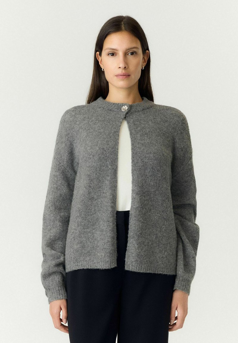 Femme aux longs cheveux foncés portant un cardigan gris boutonné par-dessus un haut blanc et un pantalon noir, debout devant un fond clair.