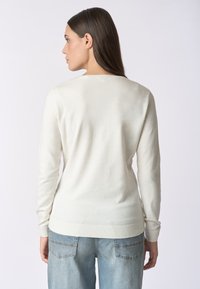Maglione crema chiaro realizzato in tessuto morbido, con scollatura rotonda, maniche lunghe e polsini e orlo a coste. Indossato con jeans azzurri chiari.