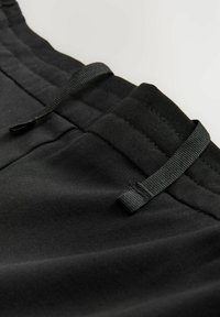 Černé sportovní kalhoty s elastickým pasem, detaily se stahovací šnurkou a texturovanou tkaninou. Detailní záběr ukazuje šití a detaily kapes.