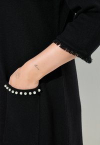 Schwarzes Strickkleidungsstück mit ausgefransten Bündchen, ausgestattet mit einer Tasche, die mit weißen Perlenakzenten verziert ist. Glatte Textur und schlankes Design.