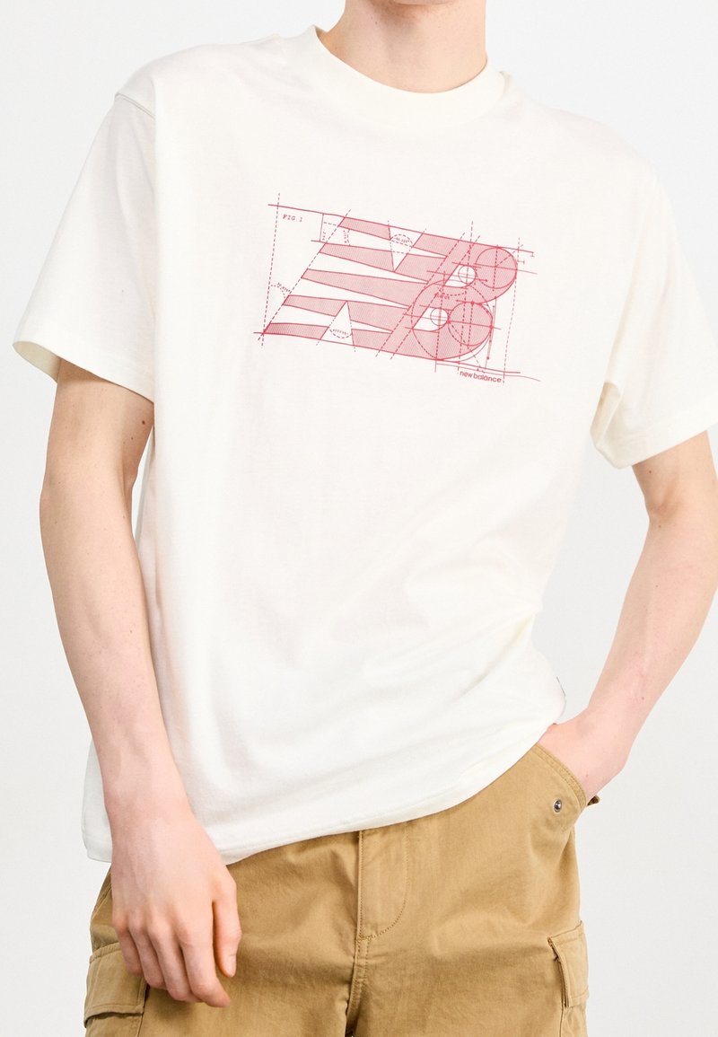Camiseta de algodón blanca de manga corta con un gráfico geométrico rojo que presenta líneas diagonales y círculos, etiquetado con "new balance" en la parte inferior.