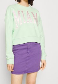 Felpa cropped verde menta con la scritta "MIAMI" in caratteri rosa lucidi, abbinata a una gonna di jeans viola con cucitura frontale e tasche.