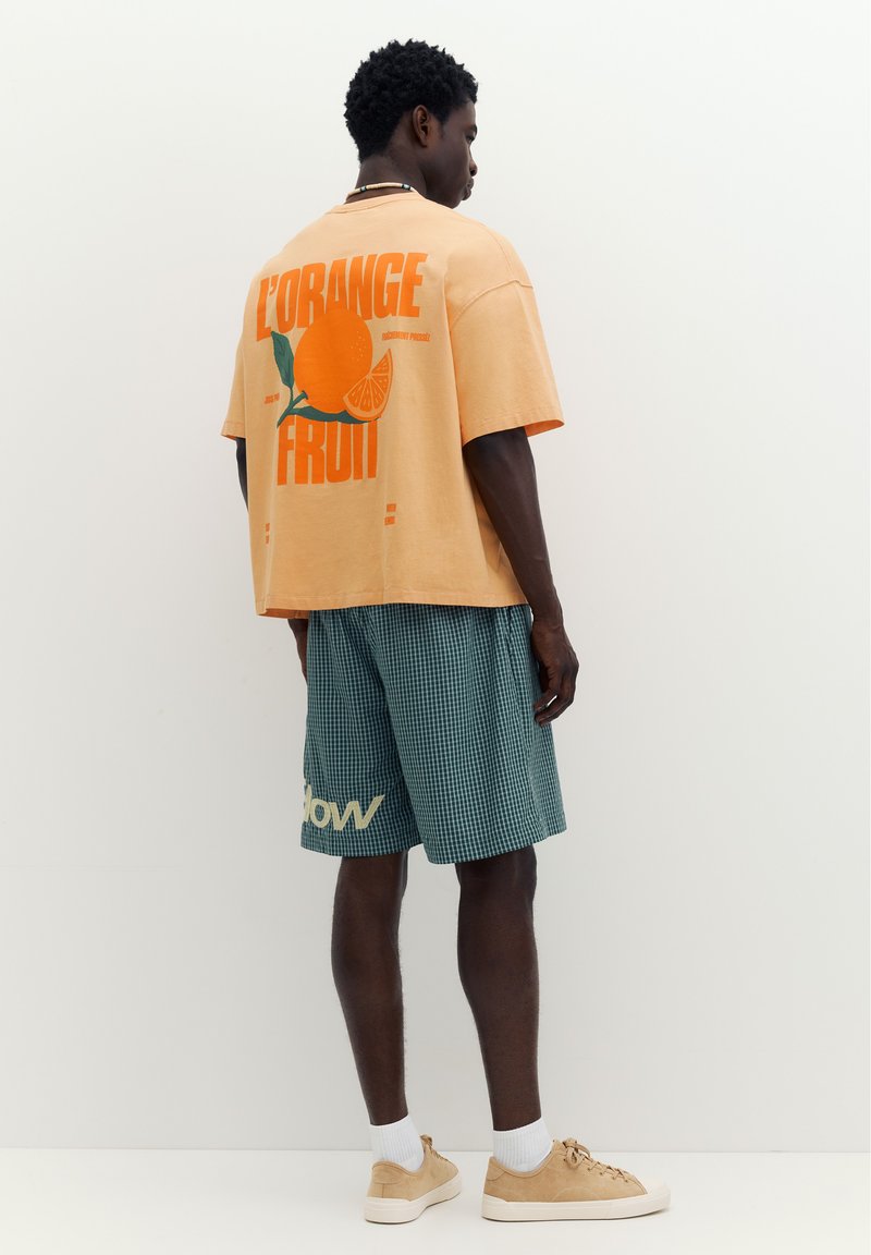 Homme portant un t-shirt oversize pêche avec un imprimé fruit orange et un short vert à carreaux avec texte, debout contre un mur blanc uni.