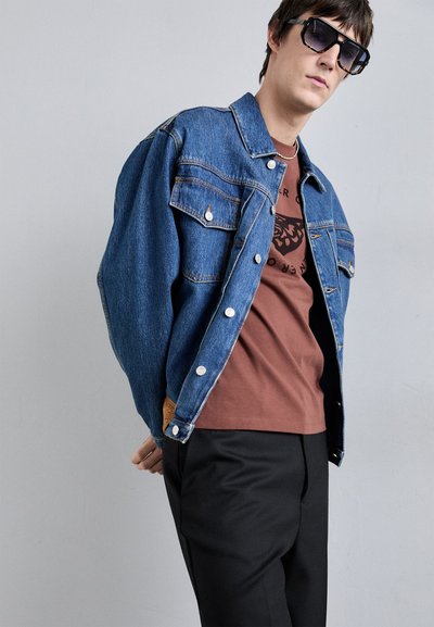 Chaqueta de denim con cuello puntiagudo, bolsillos frontales y botones plateados, llevada sobre una camiseta marrón. Pantalones negros complementan el conjunto.