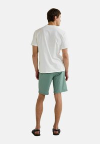 T-shirt à manches courtes blanche, shorts vert clair et sandales noires. Les vêtements sont en coton, avec un design simple sans motifs visibles.
