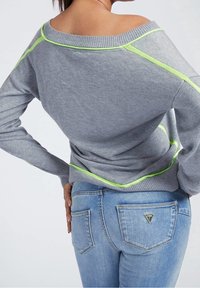 Pull gris à texture douce, design épaules dénudées avec accents vert fluo ; assorti à un jean bleu clair avec poches zippées.