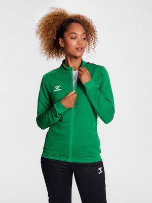 Vrouw met krullend haar die een groene hummel sportjas dichtritst, draagt zwarte hummel broek en staat voor een effen witte achtergrond.