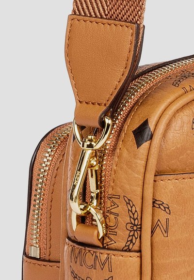 Gros plan sur le coin d'un sac en cuir brun avec un fermoir doré, une fermeture éclair et un motif noir MCM gravé sur une surface texturée.