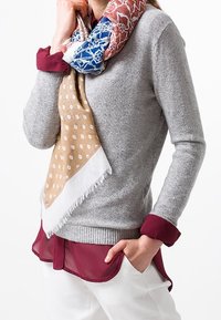 Pull gris avec bordure bordeaux, superposé à un chemisier bordeaux transparent. Une écharpe à motifs présente des tons bleus, rouges et marron avec des bords frangés.