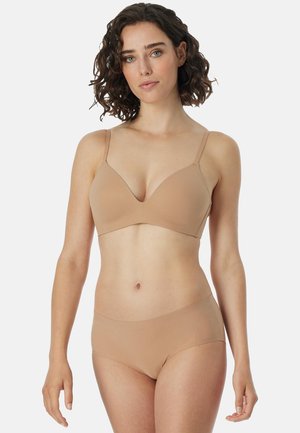Kobieta o kręconych brązowych włosach, ubrana w biustonosz w kolorze nude oraz pasujące bezszwowe majtki, stojąca na jednolitym białym tle.