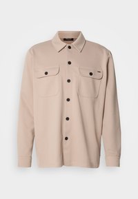 Chemise beige à manches longues avec col, boutons noirs sur le devant et deux poches poitrine boutonnées.