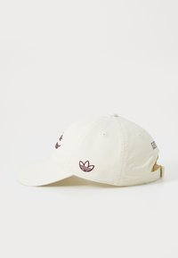 Off-white baseballcap med brodert Adidas-logo på siden og justerbar stropp bak, vist fra venstre profil.